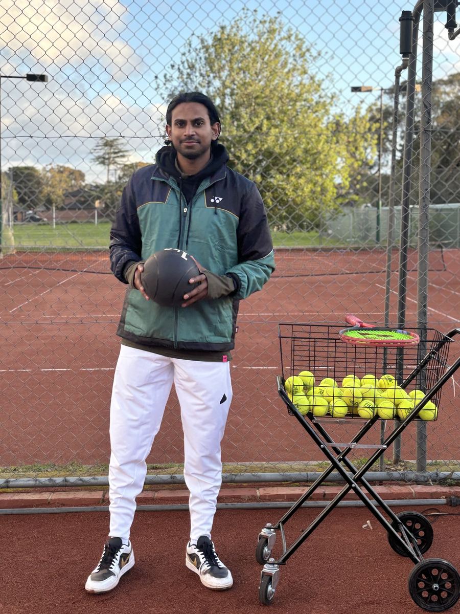 Temp Tennis coach standing on the clay courts at Holy Saviour Tennis Club, Vermont South, with a ball cart and racquets
